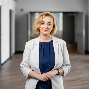 Zdjęcie przedstawia elegancką kobietę w średnim wieku z krótkimi blond włosami, ubraną w granatową bluzkę i jasnoszary żakiet. Ma subtelny uśmiech i profesjonalny, ale przyjazny wyraz twarzy. Jej postawa jest otwarta, a dłonie delikatnie splecione przed sobą. Tło ukazuje nowoczesne wnętrze z korytarzem prowadzącym do kilku pomieszczeń, co sugeruje środowisko biurowe lub instytucjonalne.