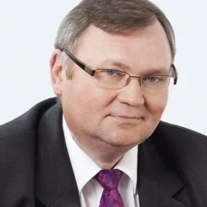 Zdjęcie przedstawia mężczyznę w średnim wieku o jasnej cerze i krótkich, ciemnych włosach. Nosi elegancki garnitur w ciemnym kolorze, białą koszulę oraz fioletowy krawat, co nadaje mu formalny i profesjonalny wygląd. Na twarzy ma subtelny uśmiech, a jego spojrzenie skierowane jest w stronę obiektywu. Okulary o prostokątnych oprawkach dodają mu powagi i inteligentnego wizerunku. Tło jest neutralne, co podkreśla jego biznesowy charakter.