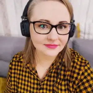 Zdjęcie przedstawia kobietę w okularach, ubraną w koszulę w czarno-żółtą kratę. Ma na sobie słuchawki nauszne, co może sugerować, że pracuje z dźwiękiem lub uczestniczy w rozmowie online. Jej lekki uśmiech i bezpośrednie spojrzenie w obiektyw nadają zdjęciu przyjazny i profesjonalny charakter. Tło jest rozmyte, co skupia uwagę na jej twarzy.