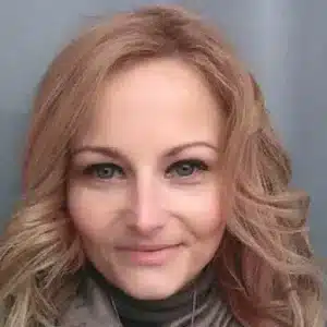 Zdjęcie przedstawia kobietę o jasnej cerze i falowanych blond włosach. Ma delikatny uśmiech i ciepłe spojrzenie skierowane prosto w obiektyw, co nadaje fotografii przyjazny i naturalny charakter. Ubrana jest w szalik i ciepłą odzież, co sugeruje, że zdjęcie mogło być wykonane w chłodniejszym sezonie. Tło jest neutralne i nie rozprasza uwagi.