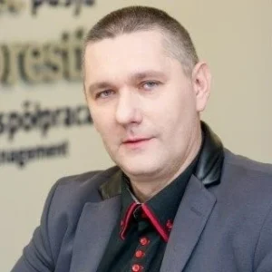 Zdjęcie przedstawia mężczyznę o jasnej cerze i krótkich, ciemnych włosach. Ma poważny, ale profesjonalny wyraz twarzy, a jego spojrzenie skierowane jest w stronę obiektywu. Ubrany jest w elegancką ciemną marynarkę z kontrastowymi detalami oraz koszulę z czerwonymi akcentami, co nadaje mu nowoczesny i biznesowy wygląd. Tło to wnętrze biurowe z widocznym napisem na ścianie, co sugeruje profesjonalne środowisko pracy.