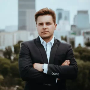 Zdjęcie przedstawia młodego mężczyznę w eleganckim garniturze, stojącego na tle panoramy miasta z nowoczesnymi wieżowcami. Ma poważny, pewny siebie wyraz twarzy i skrzyżowane ramiona, co podkreśla profesjonalny i zdecydowany wizerunek. Jego postawa sugeruje sukces i determinację.