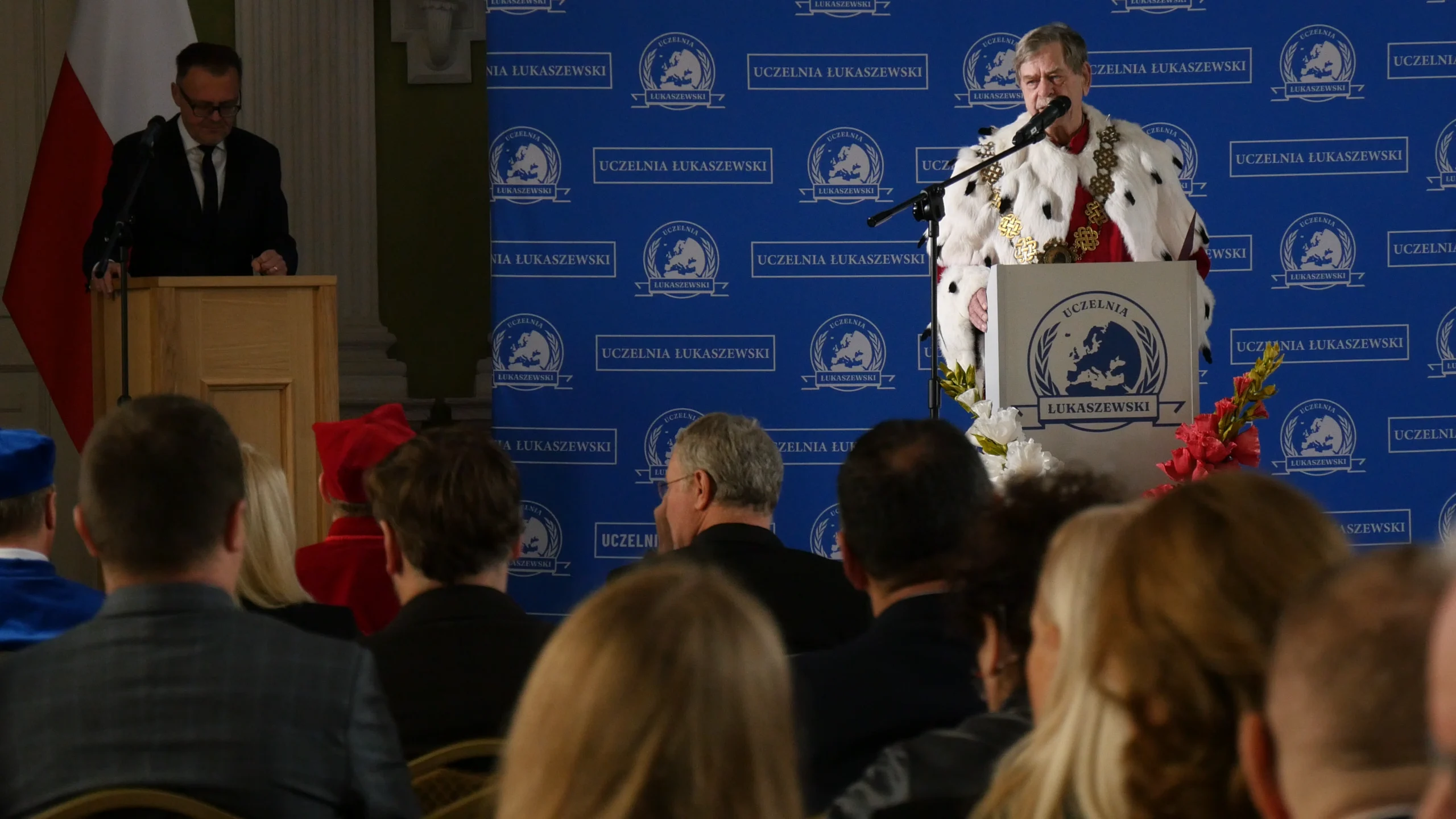 rektor uczelni podczas przemówienia na tle ścianki uczelni