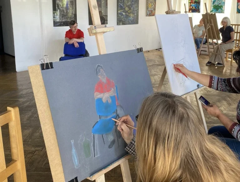 Zdjęcie przedstawia zajęcia praktyczne z malarstwa i historii sztuki na studiach podyplomowych, podczas których widoczny jest sztaluga, artysta malujący farbami, a w tle sugerująca wystawę aranżacja.